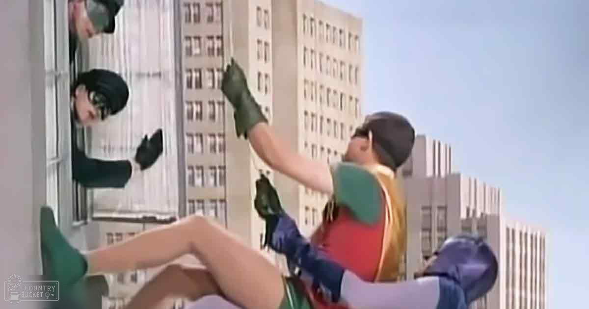 An adult female superhero in a green bodysuit and boots stands beside an adult male superhero in a red and gold chest piece, blue gloves with white accents, and green and yellow top against tall urban buildings under clear skies.