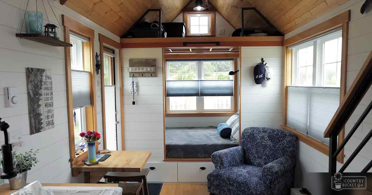 A cozy, compact living space with a wooden dining table, blue armchair, small kitchenette area, shelves holding decorative items, bed with dark mattress and pillows, indoor setting with natural light during daytime.
