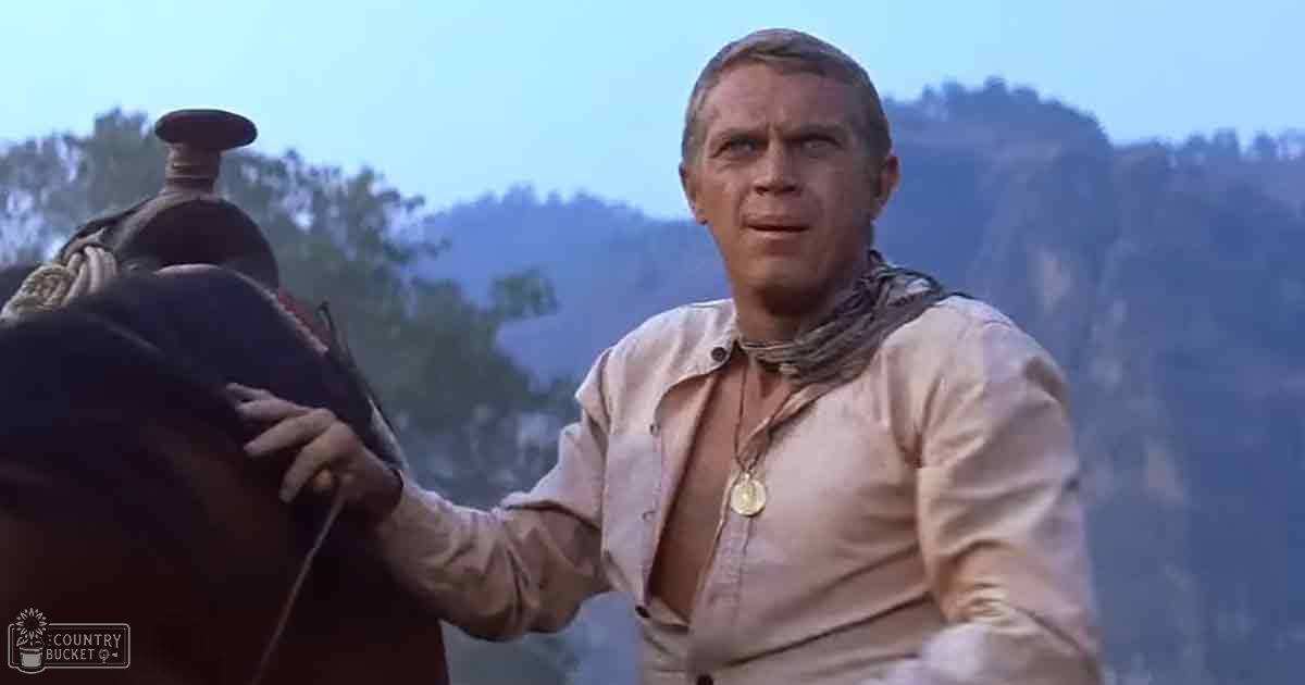 Adult male wearing a beige button-down shirt and necklace in an outdoor Western scene with mountains under clear blue sky, horse saddle visible behind him.