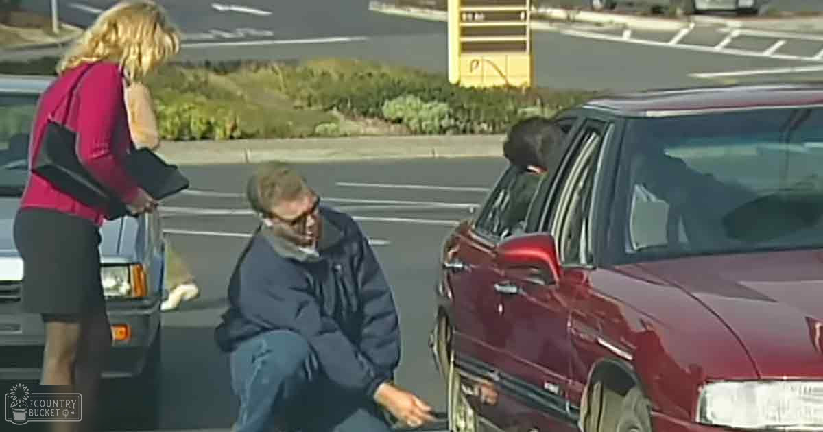An older adult male wearing a dark blue jacket and jeans stands near a maroon sedan in what appears to be a parking lot with lush greenery in the background, suggesting it might be spring or summer. A young adult female is also present, dressed in a pink top.