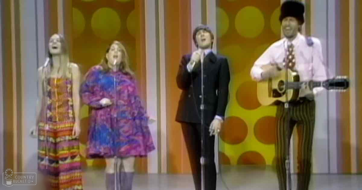 A musical performance in a studio setting with four adult performers wearing varied outfits, including striped dresses and formal suits.