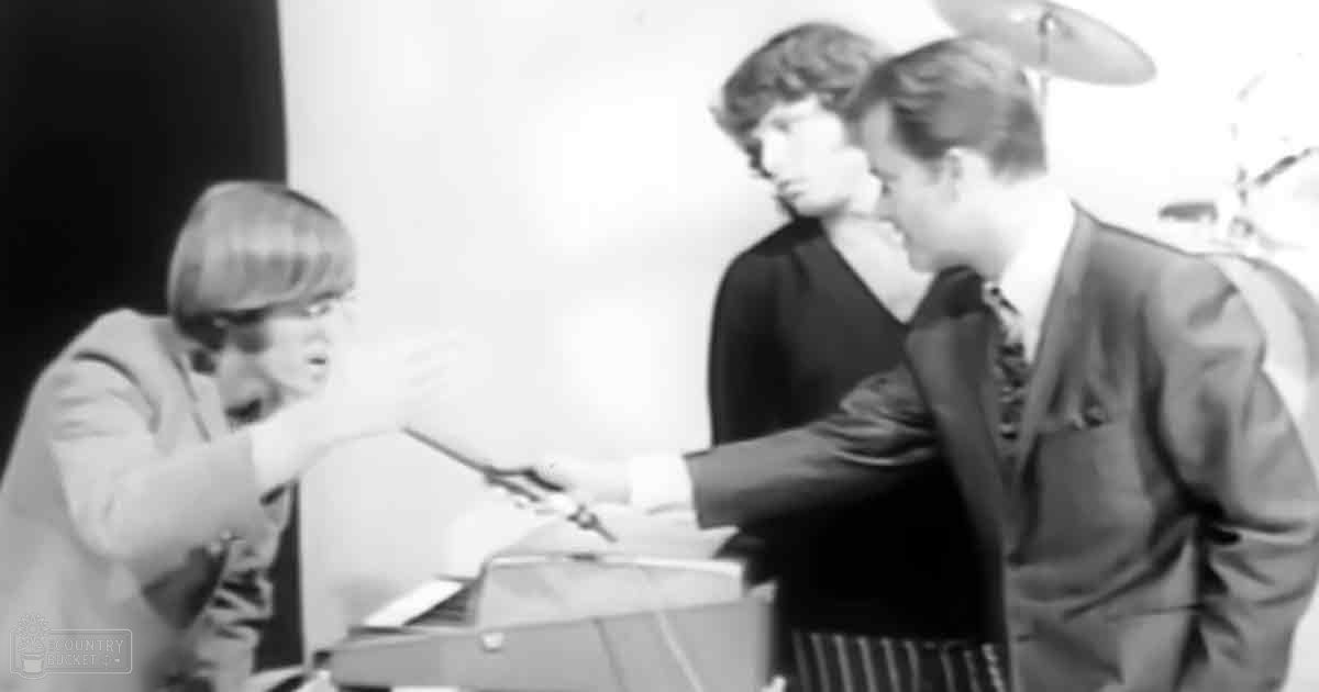 A young adult male wearing a light-colored shirt with a relaxed fit, standing next to another young adult male in a dark-colored long-sleeved shirt and tie, both indoors possibly in an office or studio setting.