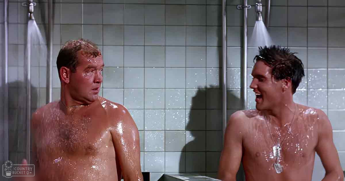 Two men in a shower setting, middle-aged and young adult males with wet skin, no upper garments, gold necklace on one, silver dog tag on another, white tiled walls, bright daylight, 'Madly Odd' watermark at bottom left.