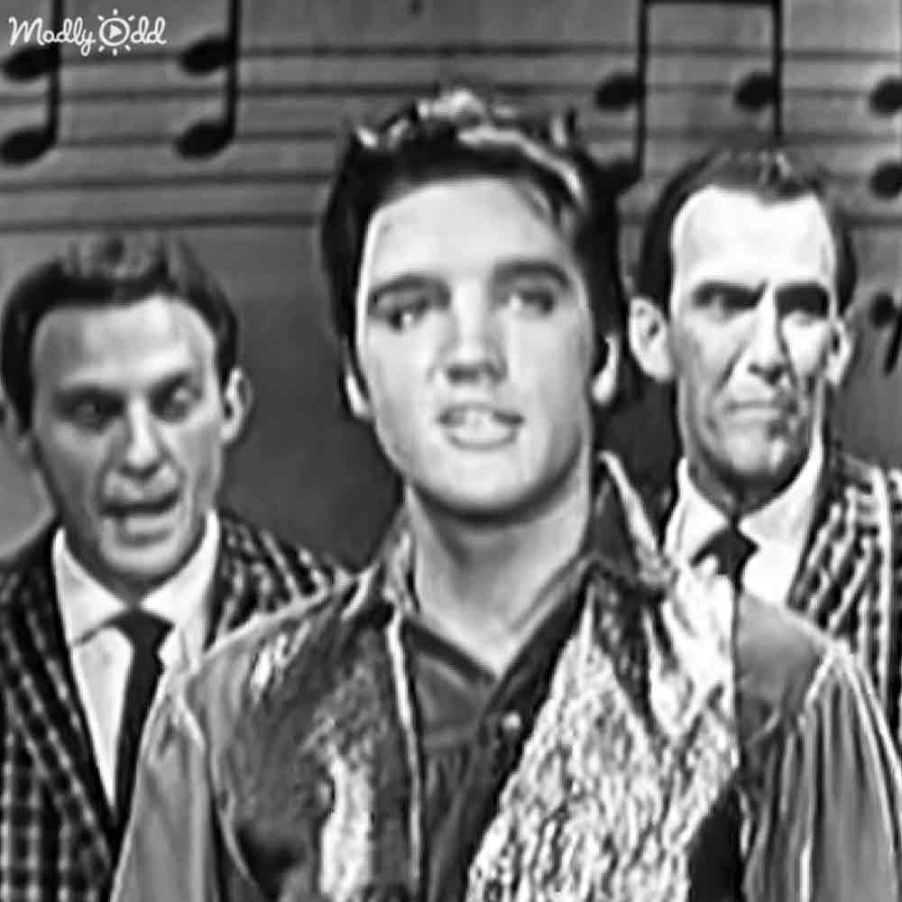 A young adult male stands at center, flanked by two other adults; a wooden wall with musical notes suggests a performance or recording studio setting.