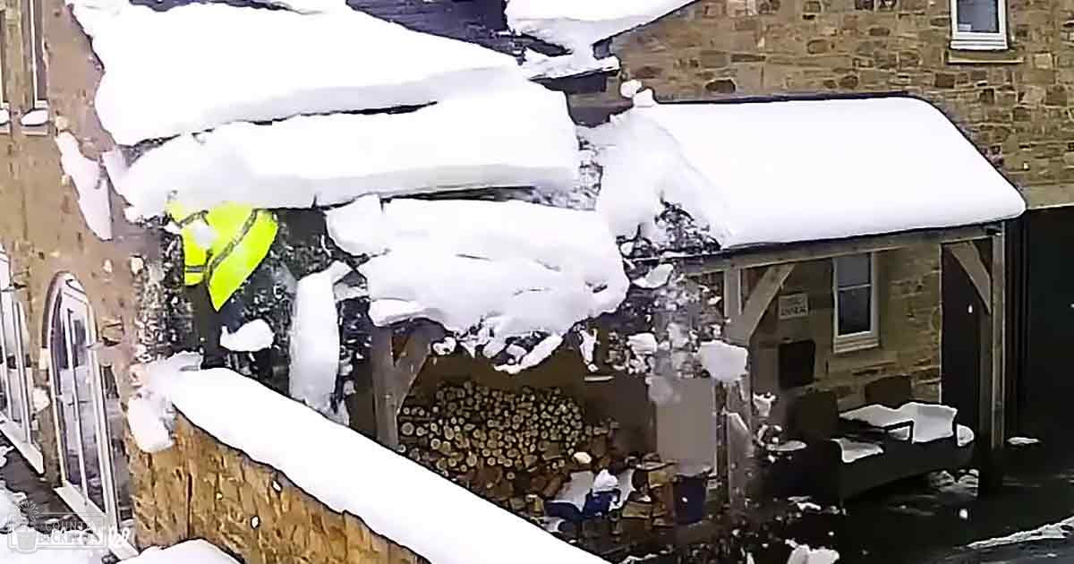 A residential winter scene with snow removal operations, a bright yellow high-visibility jacket, and heavy snowfall covering stone buildings.