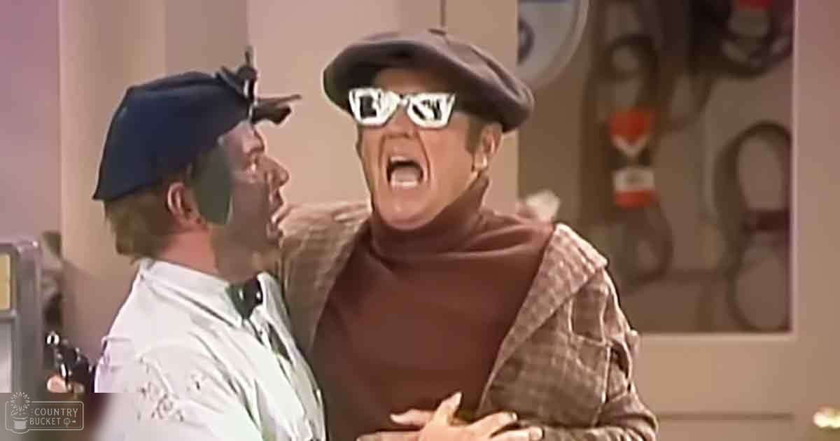 A comedic scene from a TV show or movie set in an indoor location with two men wearing casual attire. The setting includes various objects and decorations on shelves or walls behind them, suggesting a home or office environment.