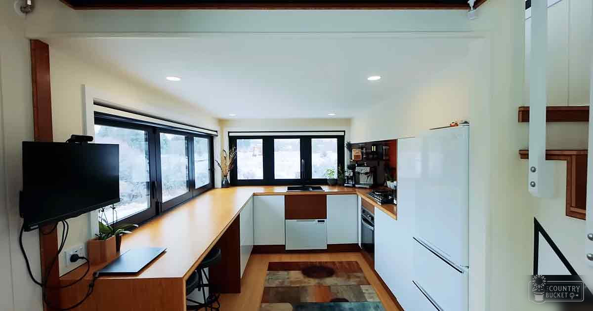A modern kitchen space with white cabinets, a wooden countertop, a flat-screen TV mounted on the wall, natural light streaming through two large windows, and a small rug in front of the stove area.