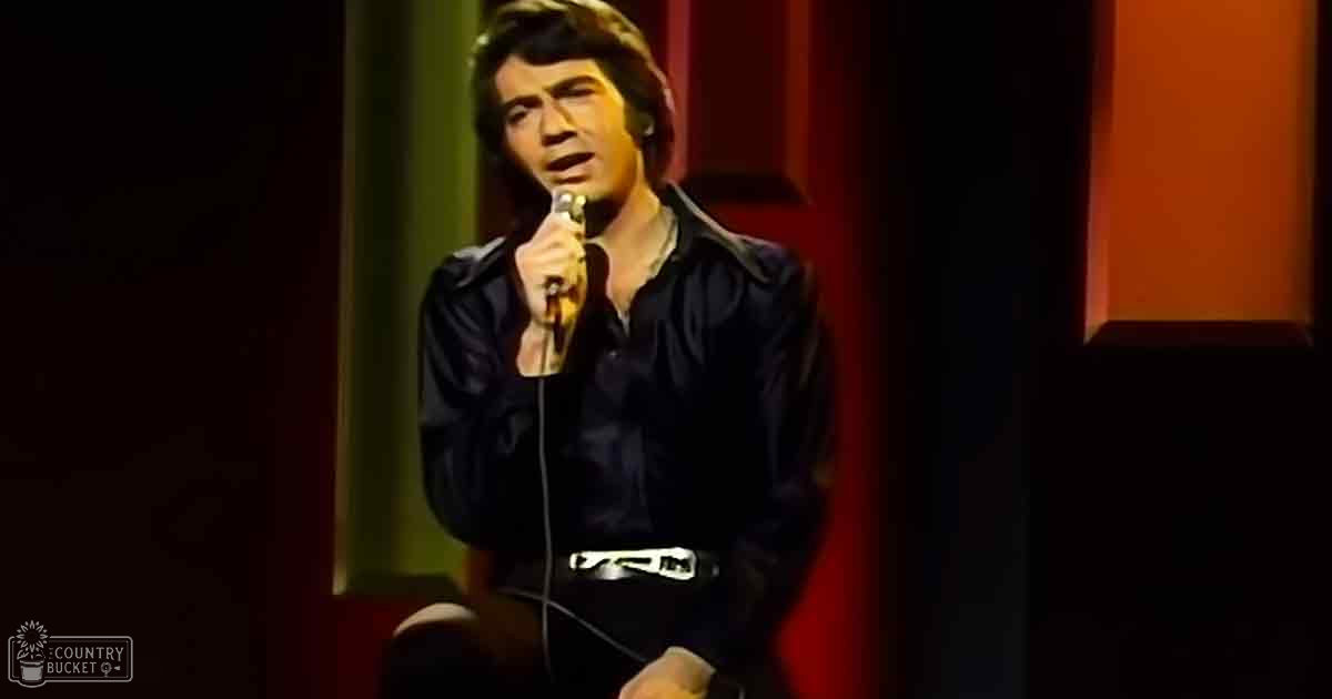 A male adult singer wearing a dark shirt, belt, and light-colored footwear, performing into a microphone in an indoor setting with red curtains.