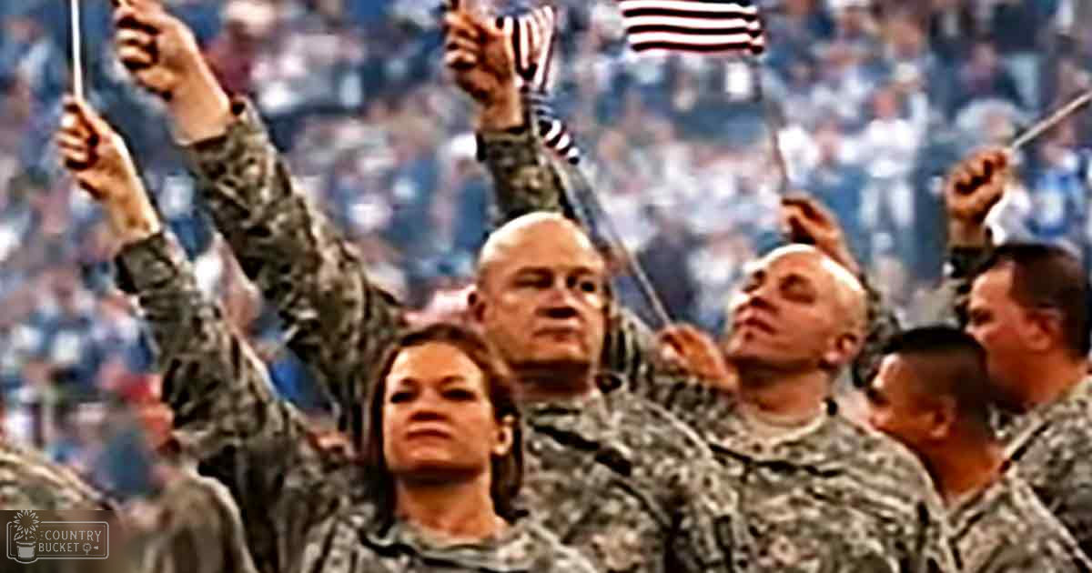 Adult female wearing camouflage-patterned military uniform at a stadium event; adult male in similar attire; group activity involving American flags under natural daylight.