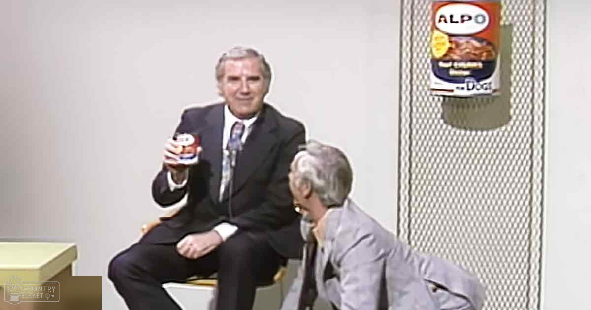 A middle-aged man in a dark suit holding an Alpo dog food can, seated indoors with another person nearby.