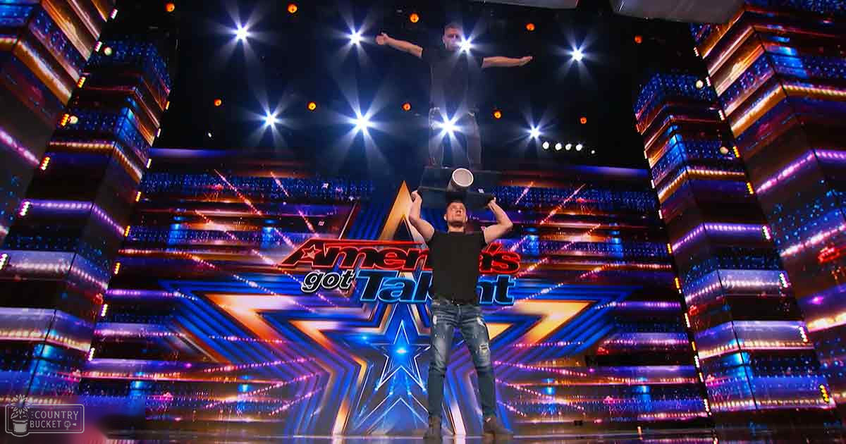 A performer balancing an object on his head at 'America's Got Talent' in front of judges, wearing a black t-shirt and blue jeans with rips.