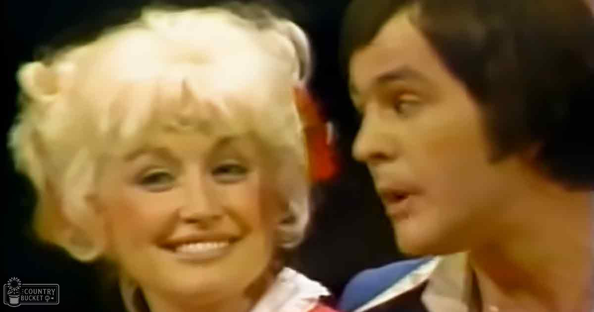 A middle-aged woman in a white dress with red accents stands beside a similarly aged man wearing a collared shirt at an indoor event or performance setting.