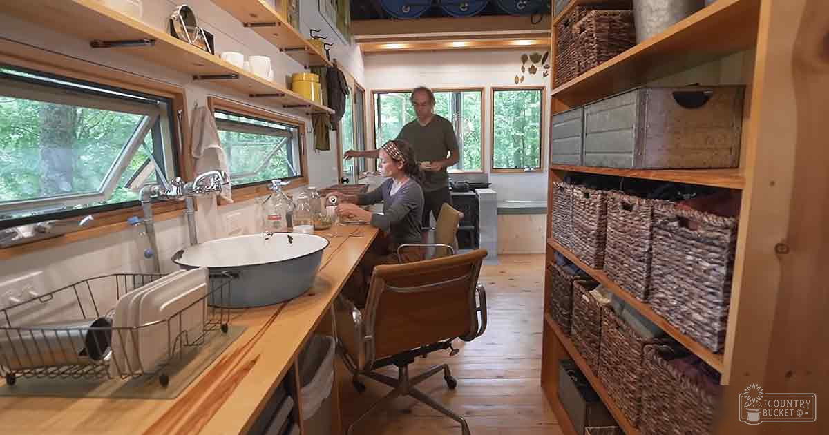 A small kitchen scene with wooden shelves filled with baskets and boxes, a sink area with natural light, and greenery outside visible through windows.