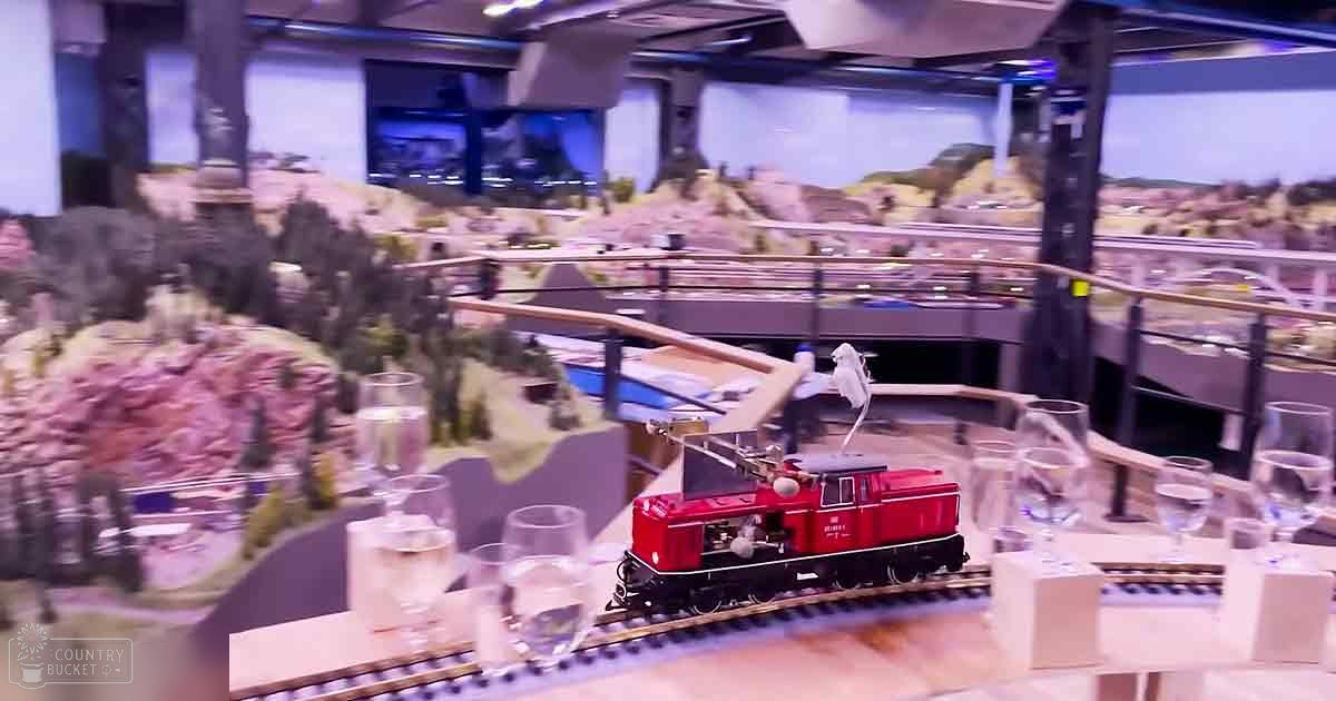A detailed miniature landscape with mountains, bridges, a red electric locomotive on tracks, and wine glasses in an indoor exhibition space.