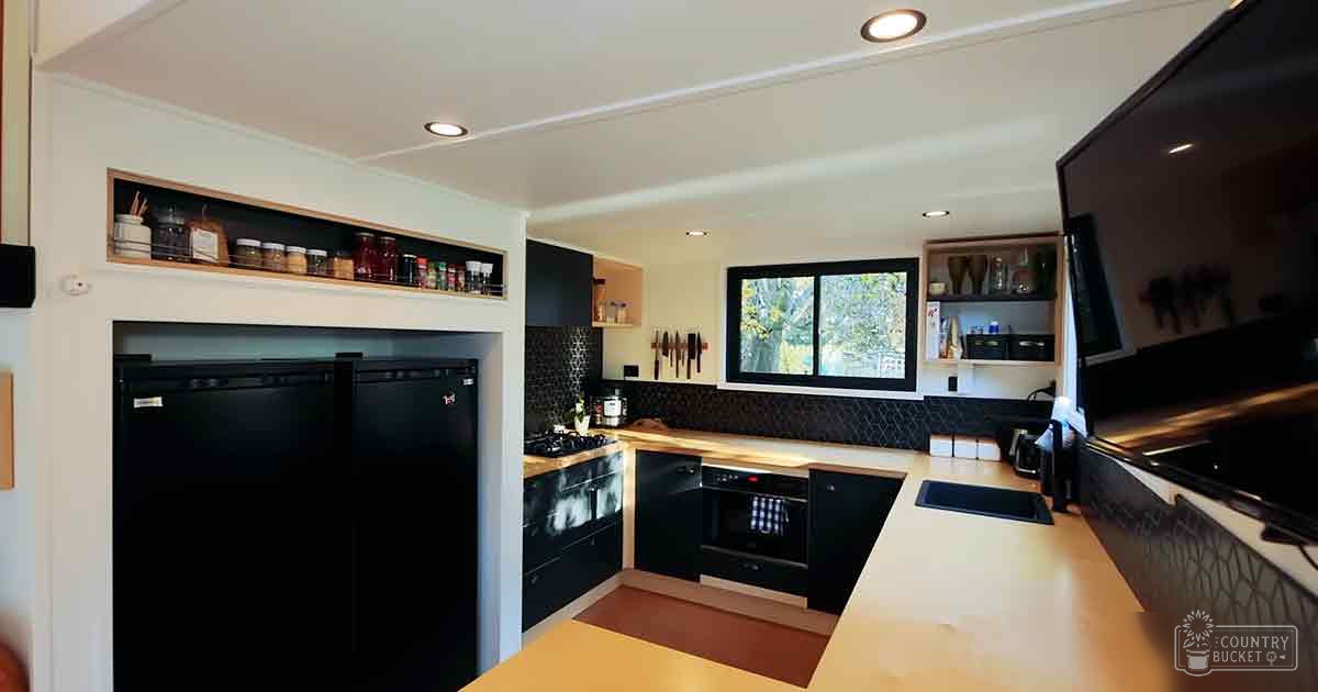 A residential kitchen with natural light streaming through a window in the center of the wall, with black tiled backsplash, open shelving holding jars and utensils, and a countertop extending along one side.