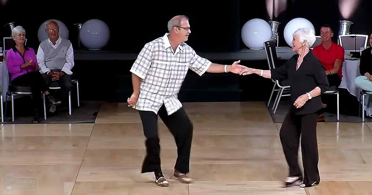 Adult male dancer wearing glasses, plaid shirt, and brown shoes in a formal dance studio with older adult female partner. Decorative elements suggest a semi-formal atmosphere.