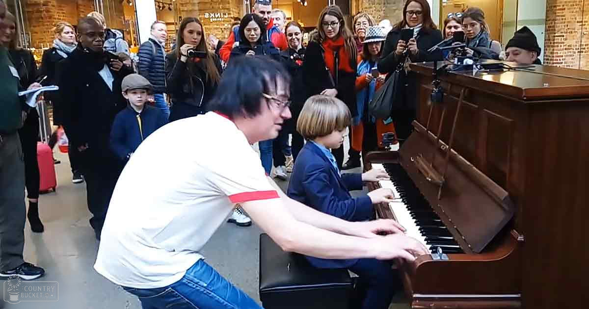 A young boy playing piano with an adult male, surrounded by diverse audience members in a public indoor space like a train station or shopping mall.