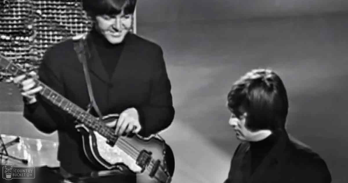 A young adult male musician plays an electric guitar indoors, wearing a dark blazer with a high collar from the 1960s era.