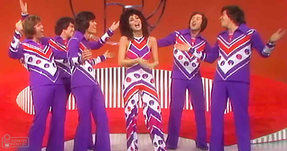 A group performing on a stage with vibrant costumes, with bold geometric patterns in purple, red, and white against a bright red backdrop.