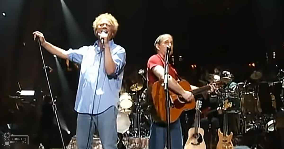 An adult male singer wearing a blue button-up shirt and light blue pants, holding a microphone stand. Next to him is another middle-aged to older adult male in a red t-shirt playing an acoustic guitar.