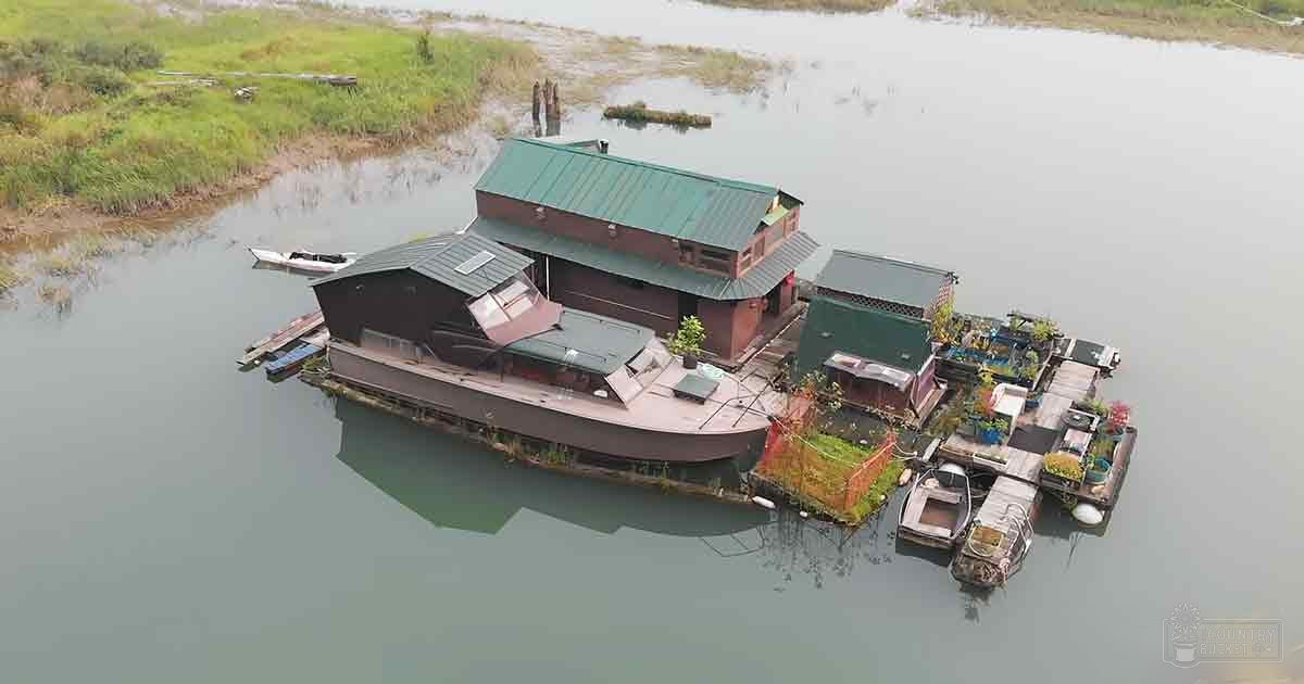 A serene rural outdoor scene with a floating houseboat surrounded by smaller boats and lush vegetation on the deck, with solar panels and potted plants visible. The setting appears to be during spring or summer.