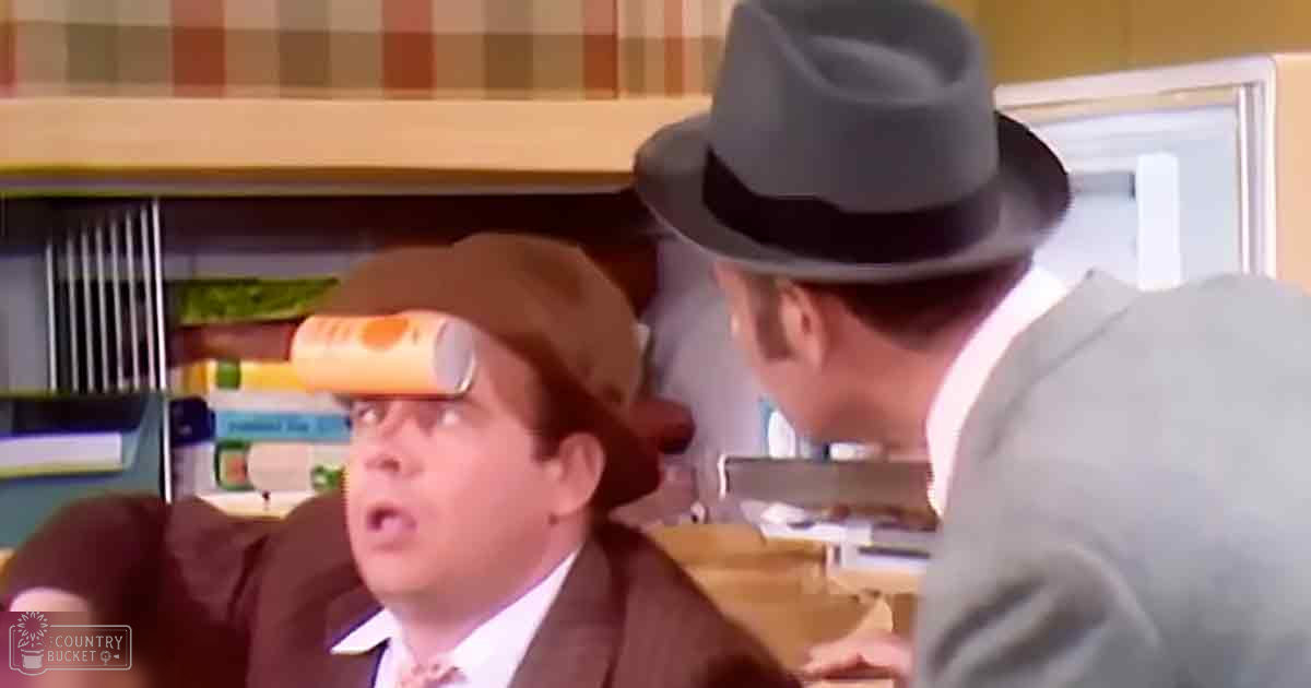 A middle-aged adult male in a brown suit jacket sits in an office, balancing a can on his backward-worn hat while another man stands nearby wearing a gray suit and fedora.