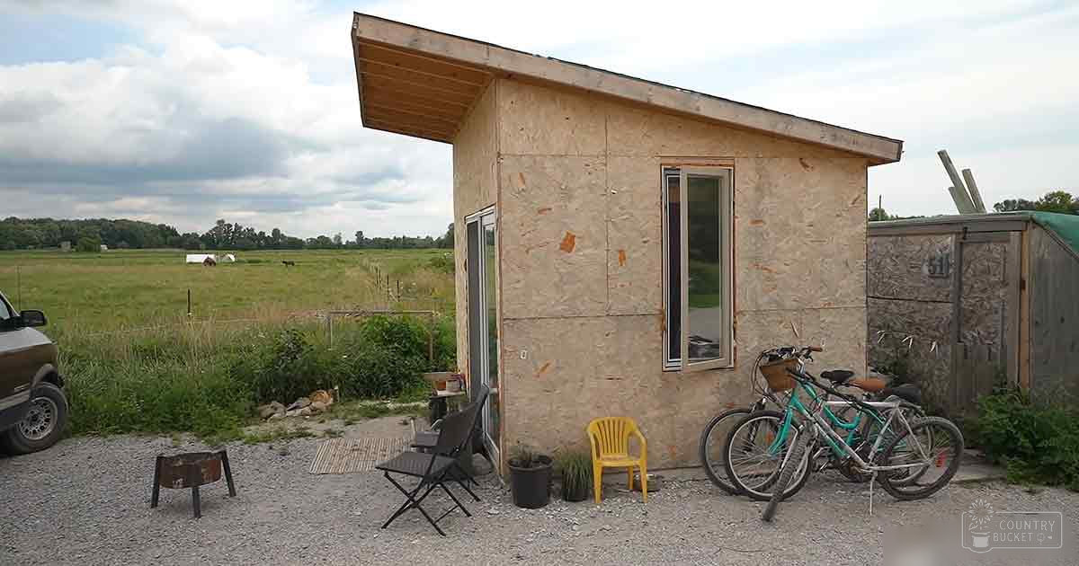 A small wooden structure in an outdoor rural setting with two bicycles, a yellow plastic chair, a black folding chair, and a potted plant near it; gravel driveway leading up to the structure under partly cloudy skies suggesting daytime spring or summer scene