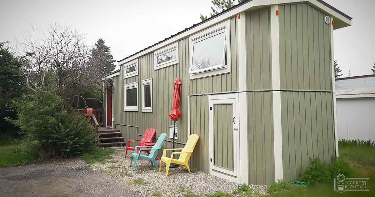 A tiny house with three colorful Adirondack chairs in an outdoor residential area with trees and overcast sky.