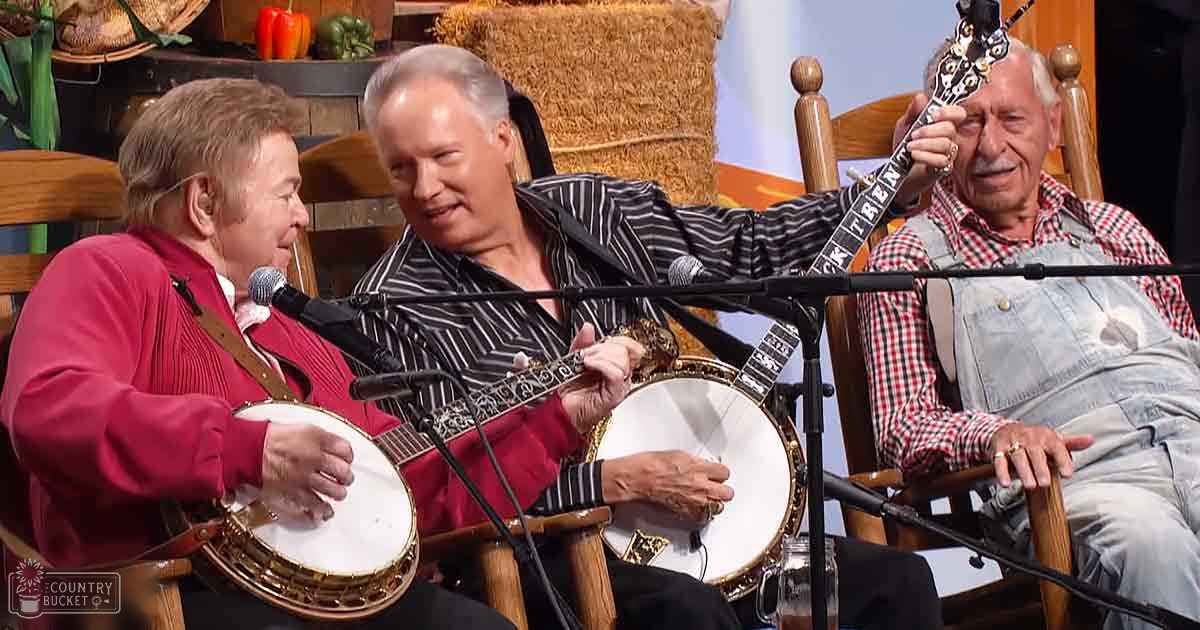 A musical performance with banjos in a rustic indoor studio or stage setup with hay bales and decorative peppers. Three adult males are present: one wearing a red sweater over a white shirt, another in a black and white striped shirt, and the third in a red and black checkered shirt with denim overalls.
