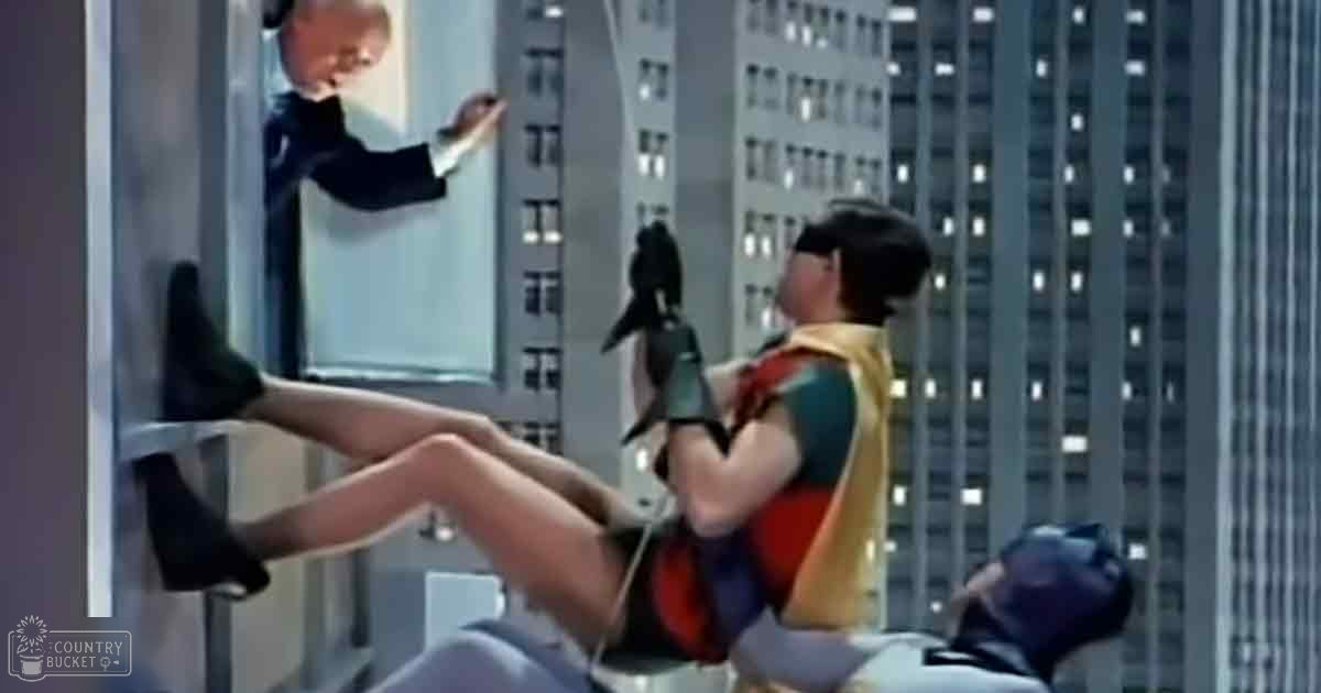 An adult male singer in a formal dark suit stands while an adult female superhero, dressed in a colorful costume with yellow and red accents, sits on a chair in front of tall urban buildings at night. A dark-furred animal with lighter patches is lying nearby.