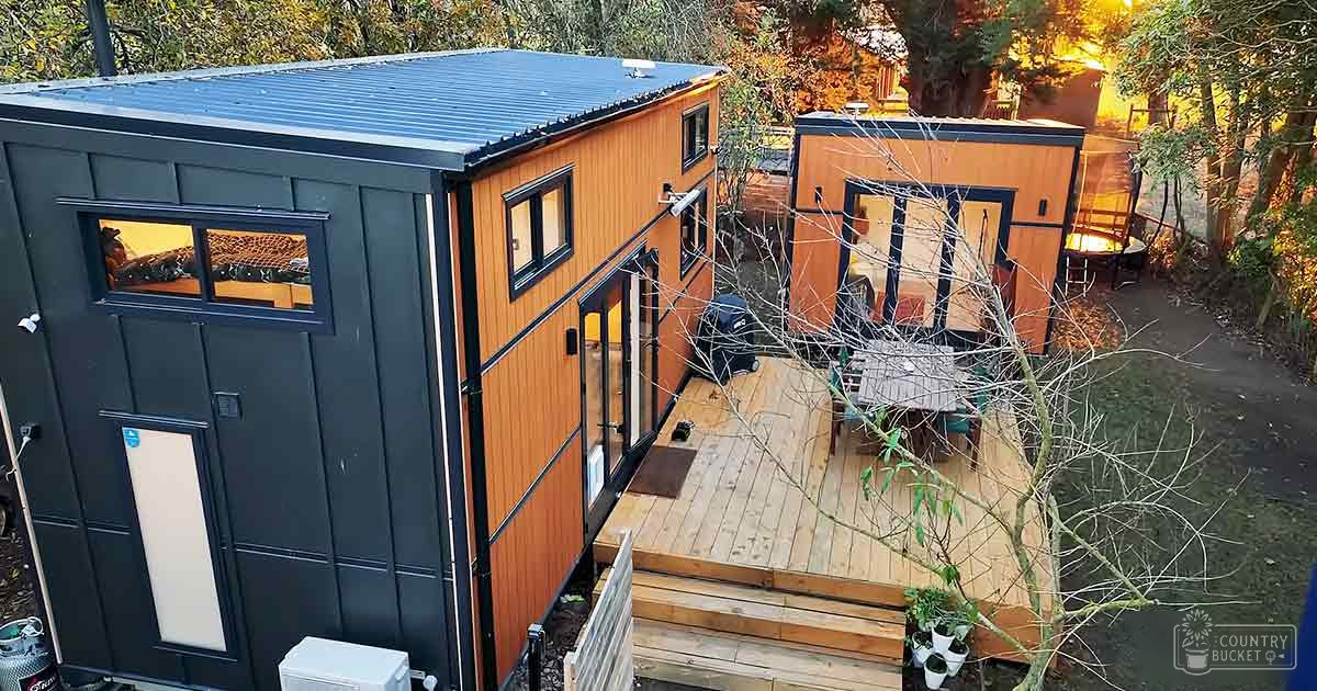 Modern tiny house community with wooden siding, black trim, outdoor furniture, potted plants, white appliance in wooded area during daylight hours.