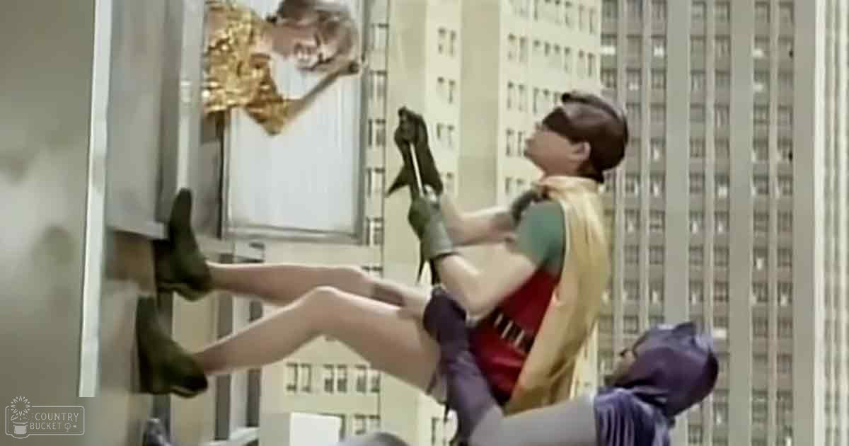 An adult male climbing the side of a tall building resembling the Empire State Building in New York City, wearing a greenish-blue t-shirt with a cape-like extension over shoulders and back, during daytime.
