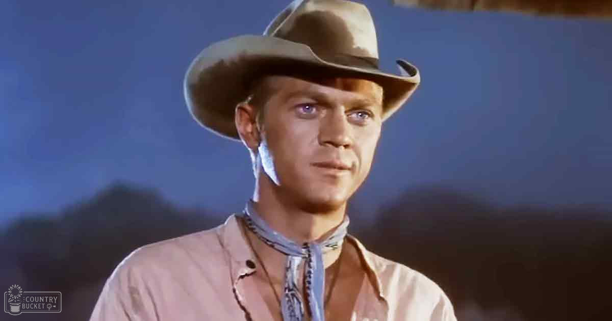 Adult male wearing a Western-style shirt with a bandana, wide-brimmed hat in an outdoor setting under dim evening light.