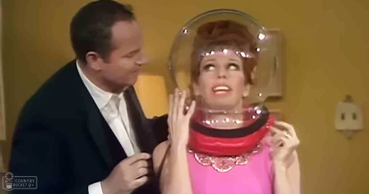 Adult male in a dark suit with white shirt and black tie, possibly mid-20th century. Adult female in bright pink dress with scalloped collar and red choker, likely also mid-20th century. Clear focus on futuristic helmet-like object worn by the woman.