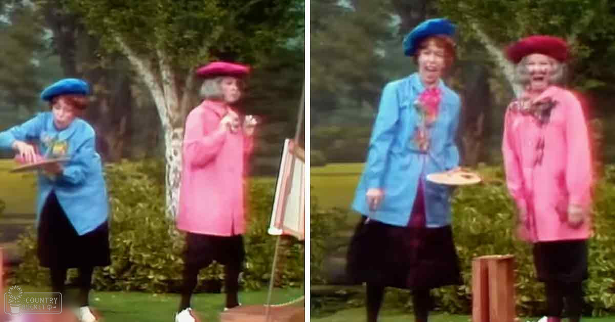 Middle-aged female performers wearing berets and coats, holding a small table with candles and a cake, set against an outdoor garden backdrop during late afternoon or early evening.