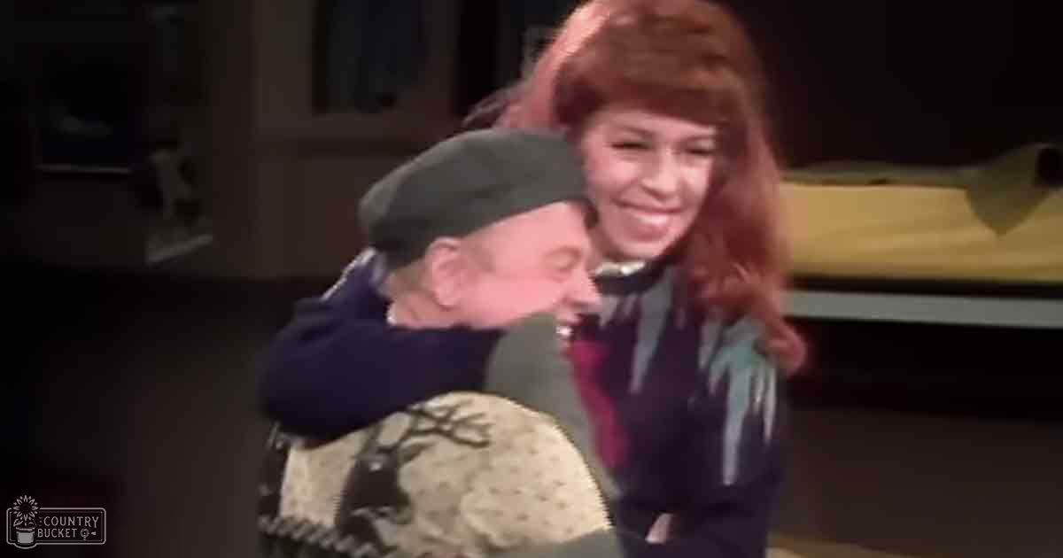 An adult female in a home setting interacting with an older male wearing a beret-style hat and a sweater with tree branch patterns, surrounded by indoor furnishings under artificial evening lighting.