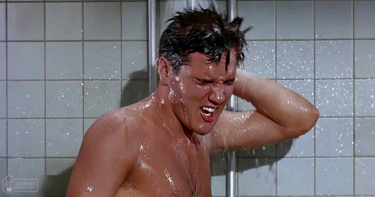 A man taking a shower with visible muscle tone under a tank top; necklace around his neck.