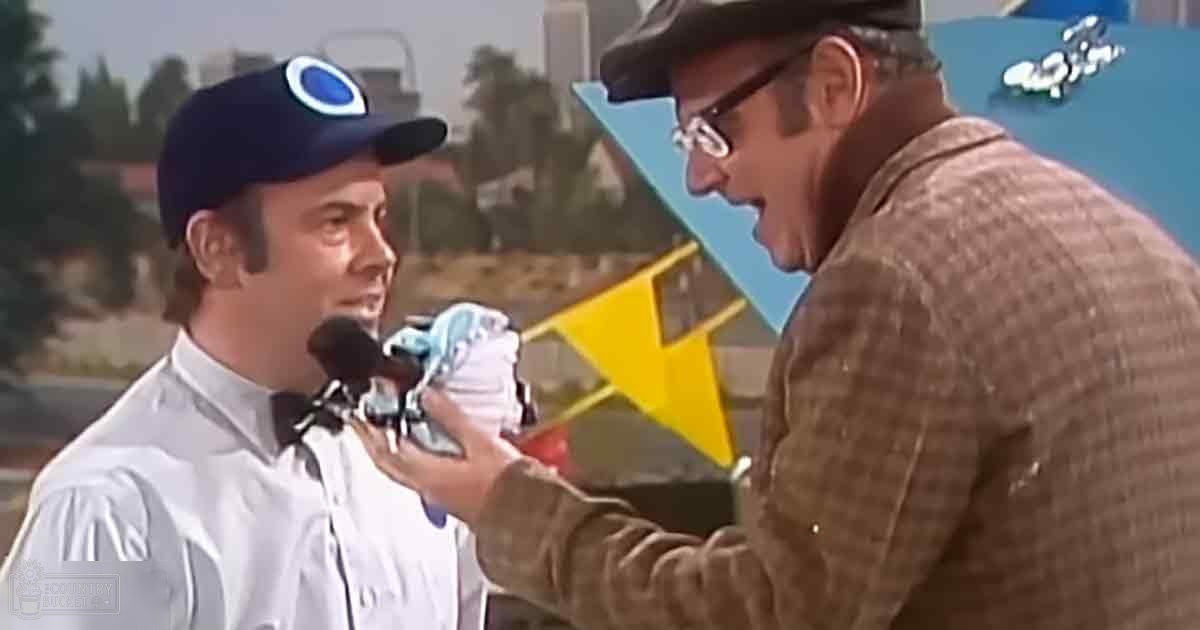 An adult male race car driver in a white racing suit being interviewed by another older adult male at a racetrack surrounded by palm trees and buildings on a clear, sunny day.