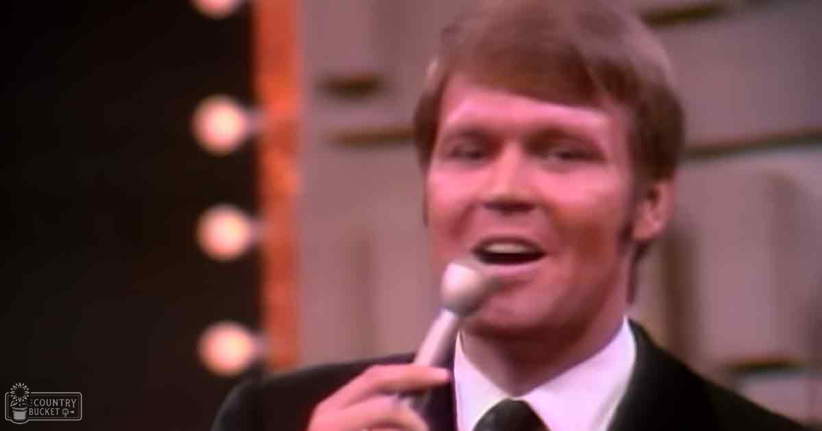 An adult male singer wearing a dark suit jacket over a white dress shirt with a dark tie, set against stage lighting suggesting a performance or broadcast from the mid-20th century.