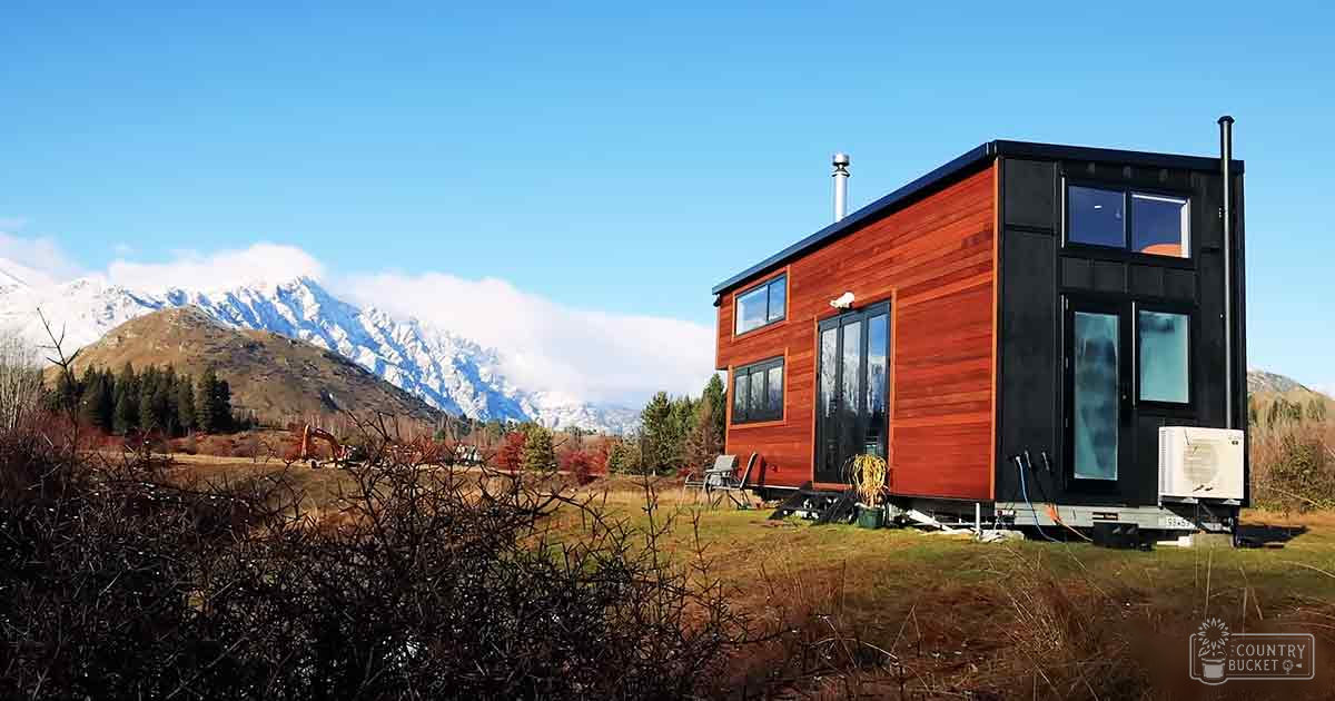 A modern tiny house with red and black exterior walls, large windows, a small outdoor chair, air conditioning unit, chimney stack in Queenstown's rural area surrounded by snow-capped mountains and autumn-colored trees under natural daylight.
