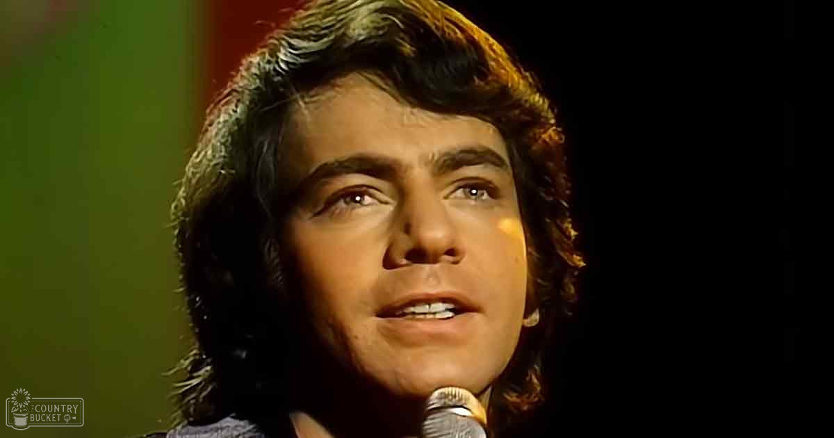 A young adult male singing into a microphone indoors under stage lighting.
