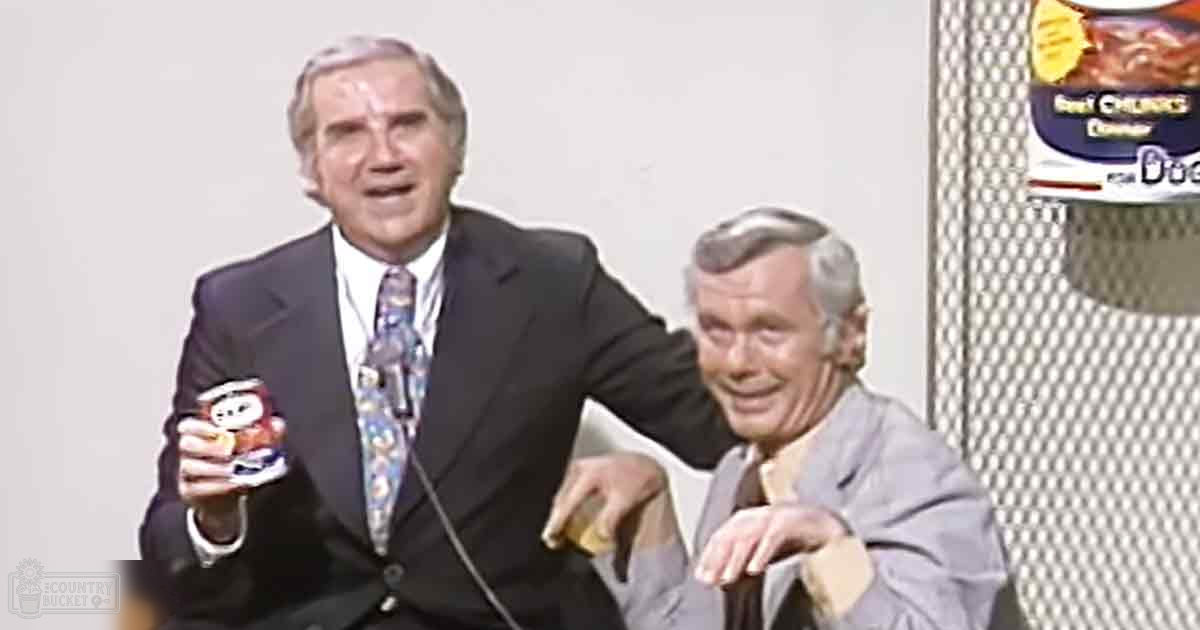 An older adult male in a dark suit with a patterned tie holds a small object, while another older man in a light gray suit leans on a desk in a studio setting.