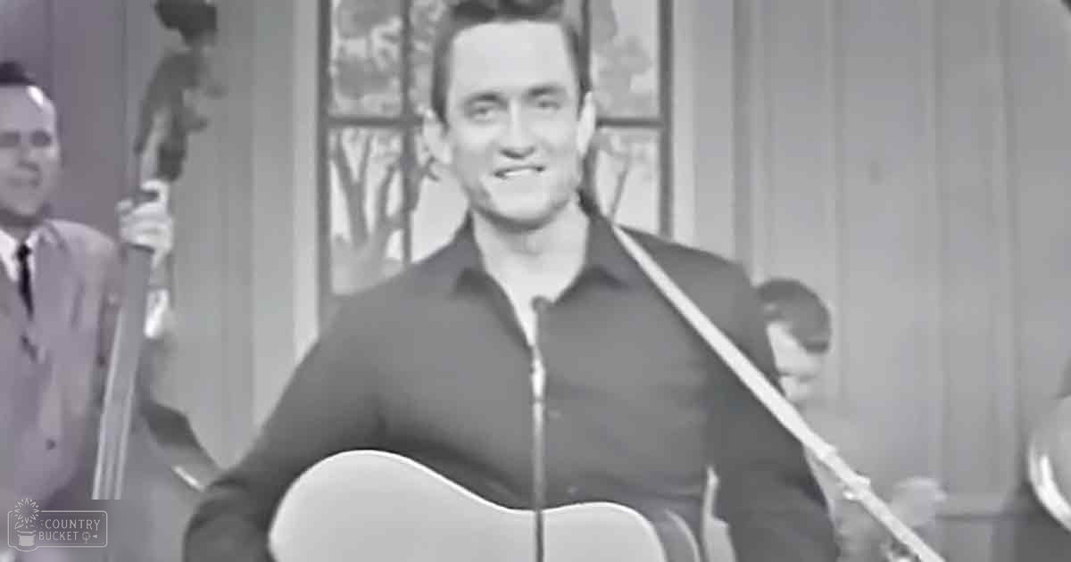 A young adult male playing guitar indoors, wearing a black zip-up shirt and a light-colored suit jacket with formal trousers.