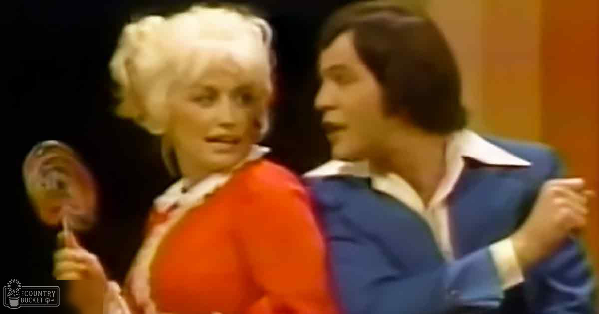 An adult female in an orange vintage-style dress holds a lollipop, accompanied by an adult male wearing a blue suit jacket and white shirt at what appears to be a performance venue with dark curtains. The scene suggests a mid-20th century setting with warm lighting.