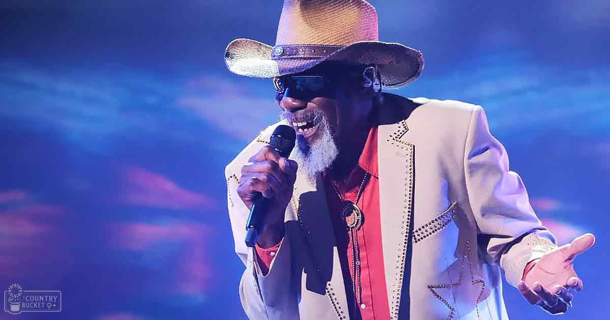 An older adult male performer wearing a white jacket adorned with gold studs, dark sunglasses, and a wide-brimmed straw hat, holding a microphone on stage under blue lighting.