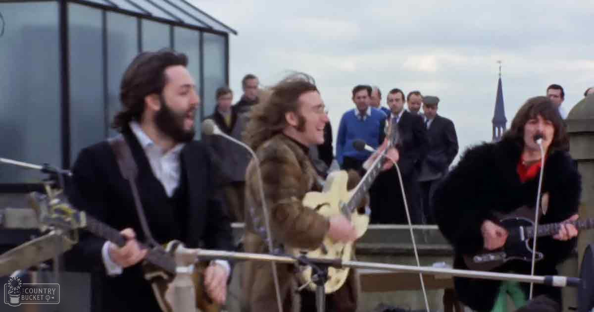 The Beatles performing their iconic rooftop concert atop Apple Corps Ltd., London, winter evening scene with urban skyline and church steeple in background.