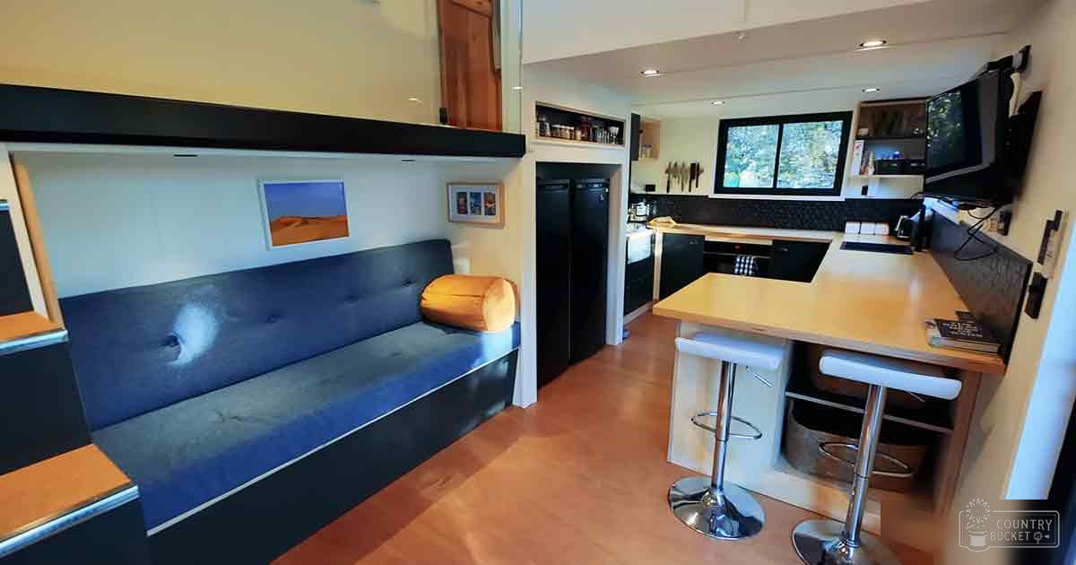 Compact living space with blue cushioned bench, wooden table, black appliances, and wall-mounted shelves in an indoor setting.
