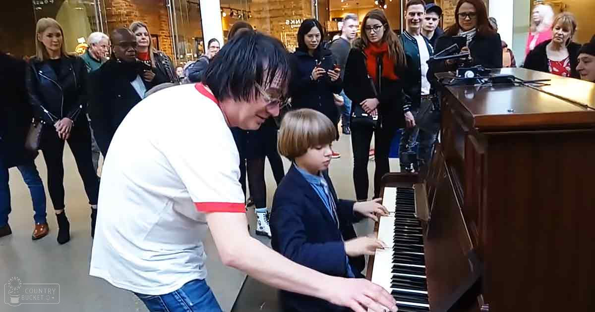 A young boy wearing a dark blue blazer and tie plays piano with an adult male in a public indoor setting, possibly a shopping mall.