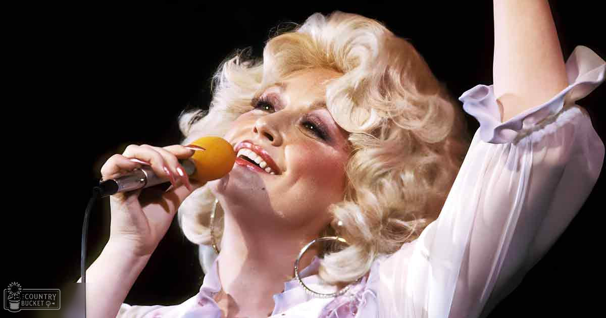 An adult female singer in a vintage-style white ruffled sleeveless dress, holding an orange-tipped microphone and wearing large hoop earrings and a bracelet, performing on stage with dimmed background lighting.