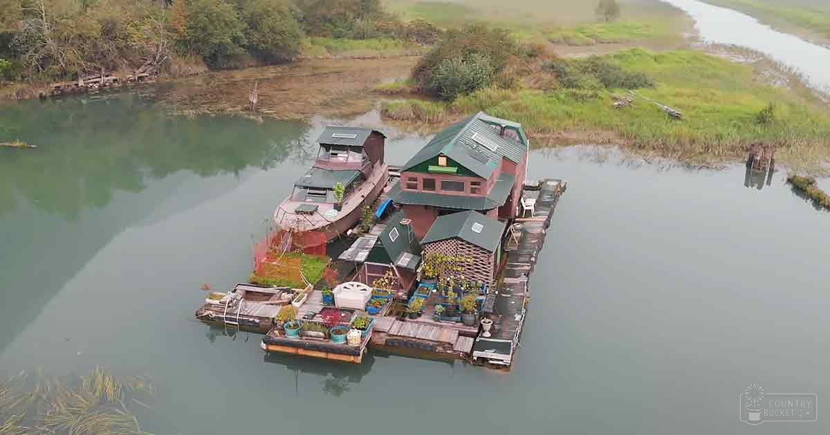 Serene floating house community surrounded by lush greenery with small boats and docks in a calm waterway bordered by dense vegetation on an overcast day, suggesting spring or summer.